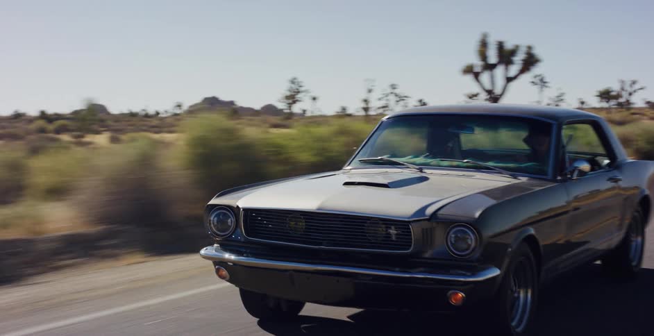 A classic grey Ford Mustang cruises down a winding desert road through Joshua Tree National Park on a sunny day