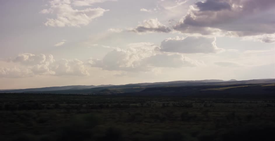 A wide shot from a moving vehicle captures a vast plain and distant hills under a cloudy sky at dusk