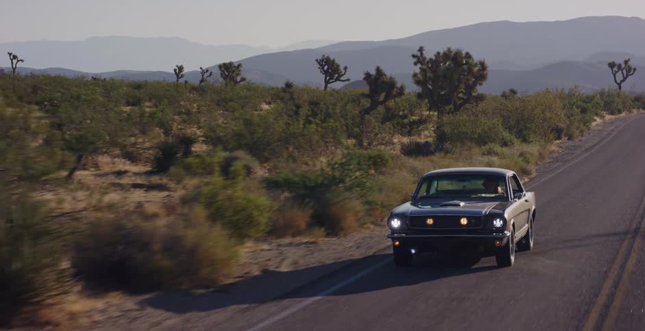 A classic black muscle car cruises down a desert highway surrounded by Joshua trees and distant mountains