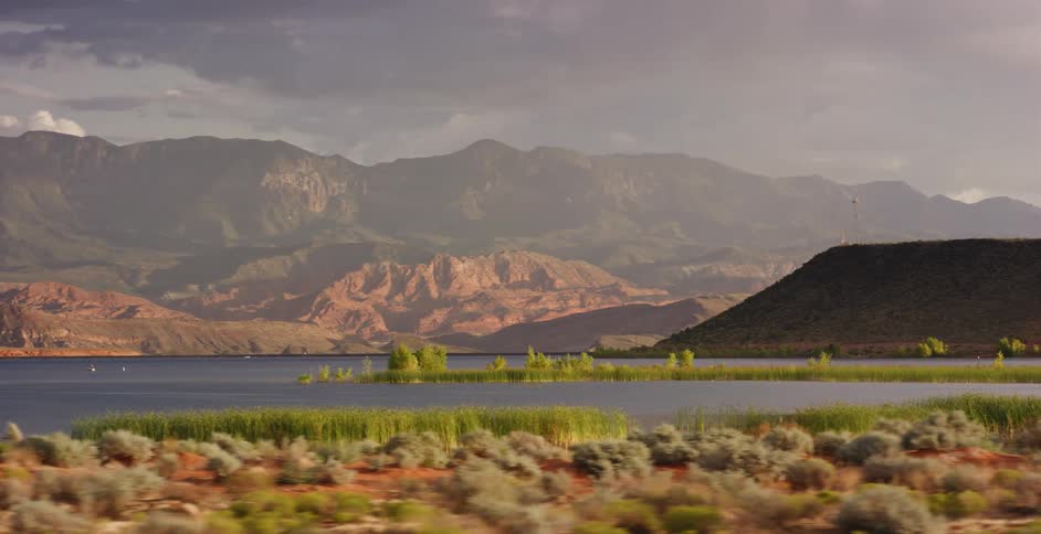 A scenic tracking shot of a desert lake surrounded by red rock mountains under a dramatic, cloudy sky
