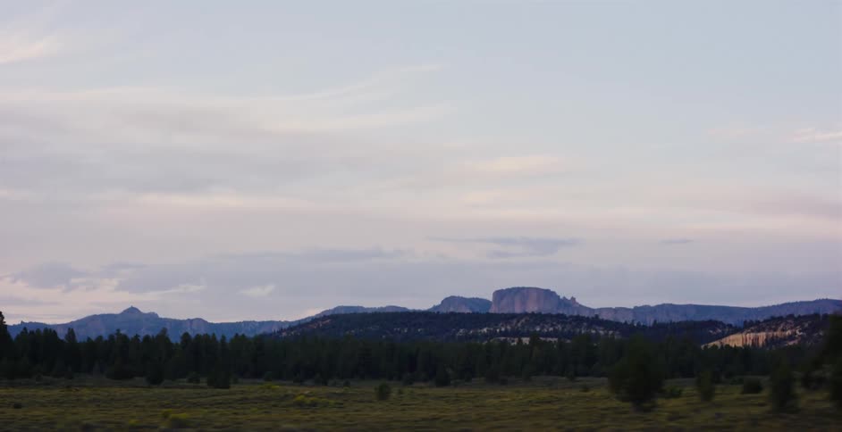 A point-of-view driving shot reveals a serene landscape of forests and distant mesas under a cloudy sky