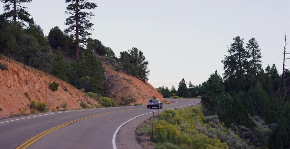 A classic car drives along a winding scenic road through a mountainous forest landscape at dusk