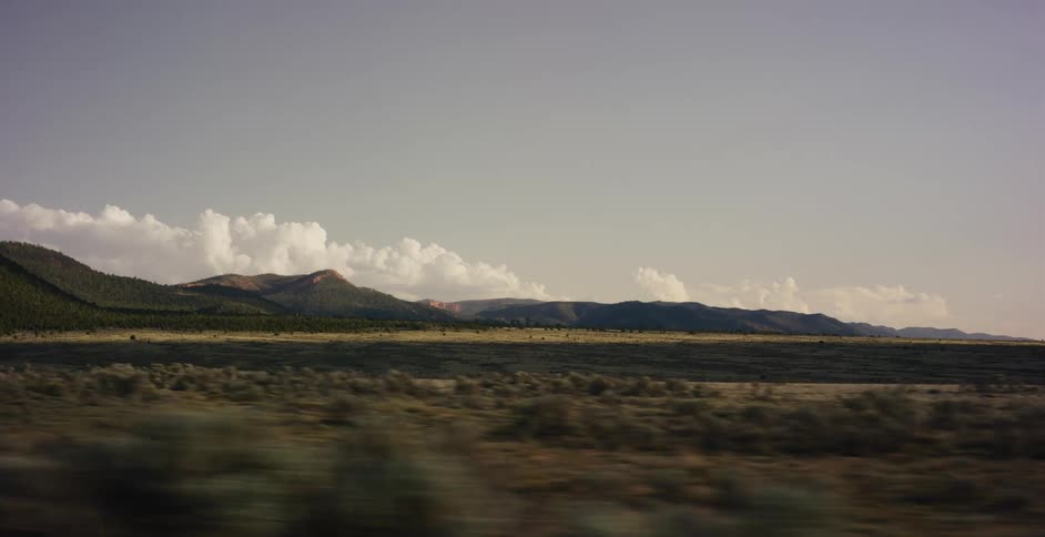 A scenic view from a moving vehicle captures a sweeping desert landscape with distant mountains under a hazy sky