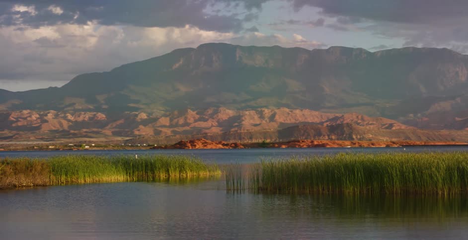 A serene wide shot of a tranquil lake with reeds zooms out to reveal a majestic red rock and mountain landscape under a partly cloudy sky