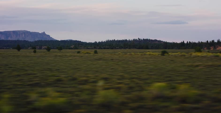 A side-view tracking shot from a moving vehicle captures a vast, green rural landscape with distant mountains at dusk