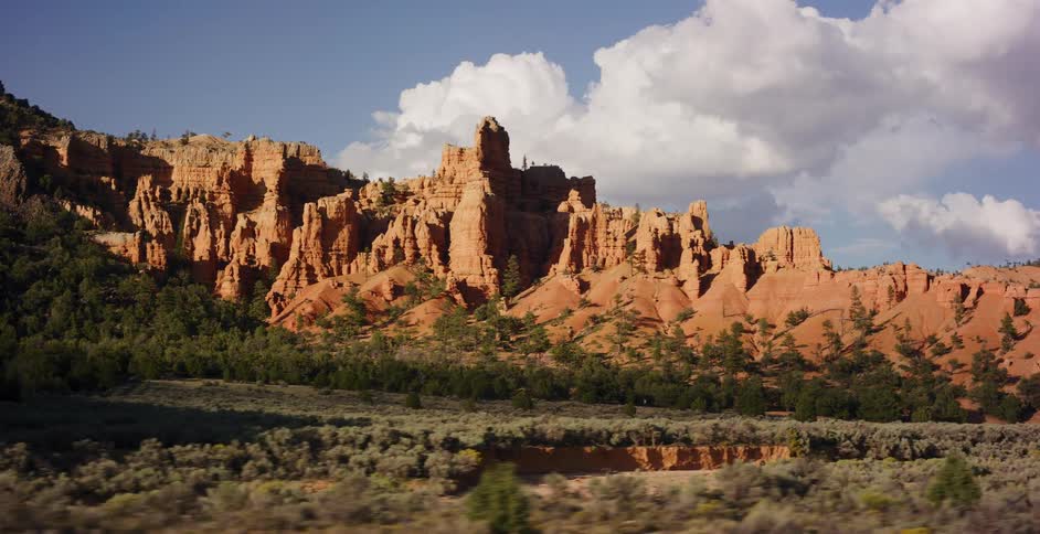 A scenic tracking shot reveals the majestic red rock formations of a desert canyon under a partly cloudy blue sky