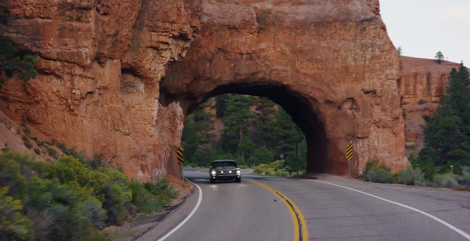 A classic black Mustang drives down a scenic highway and enters a tunnel carved through the red rock canyon