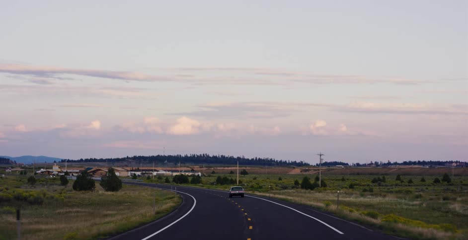 A car drives along a winding country road through an open landscape during a peaceful sunset