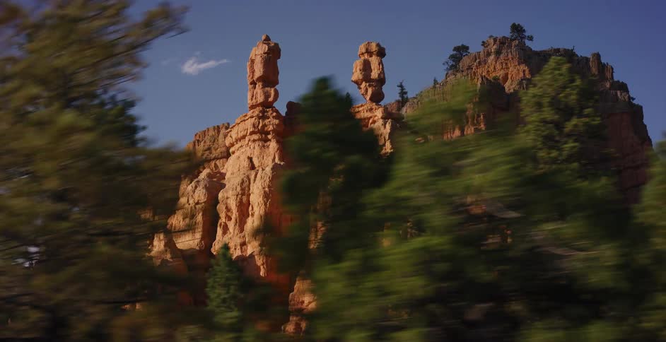 A cinematic tracking shot reveals majestic red rock hoodoo formations through a blur of green pine trees on a sunny day