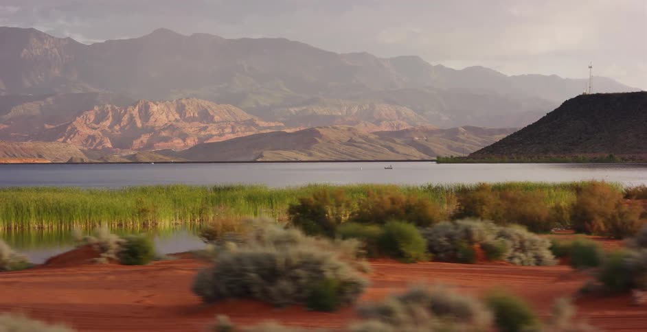 A scenic tracking shot of a tranquil lake bordered by red sand and reeds, with a stunning backdrop of desert mountains