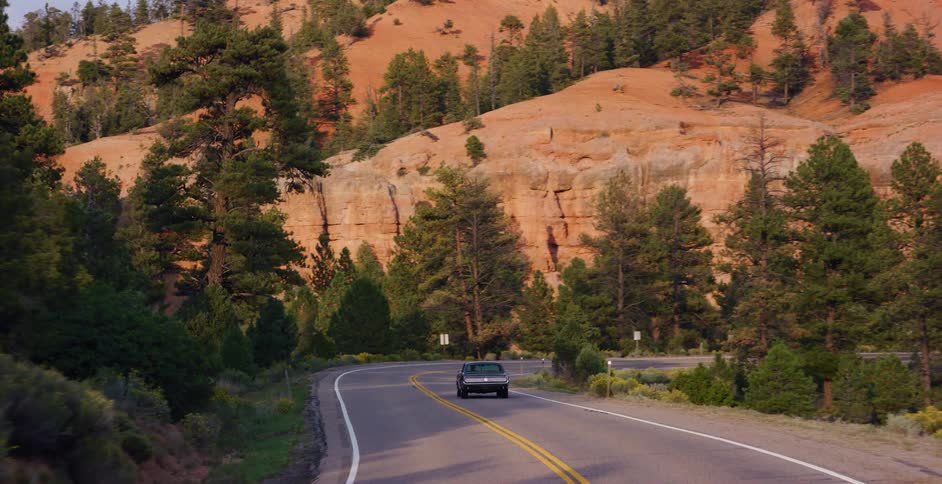 A classic car drives on a scenic, winding mountain road through a red rock canyon surrounded by pine trees