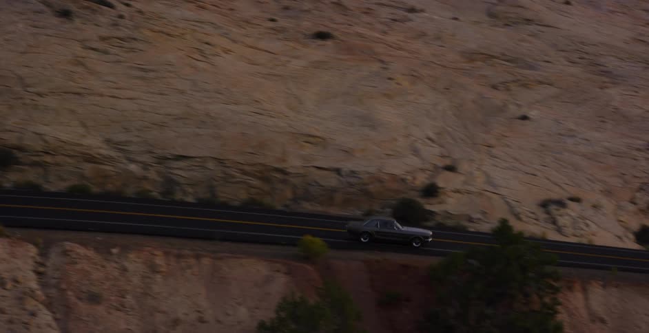An aerial view shows a classic silver muscle car driving along a scenic highway through a rocky desert canyon landscape at dusk