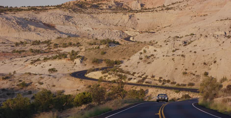 A lone car navigates the twisting turns of a desert highway surrounded by sunlit sandstone hills