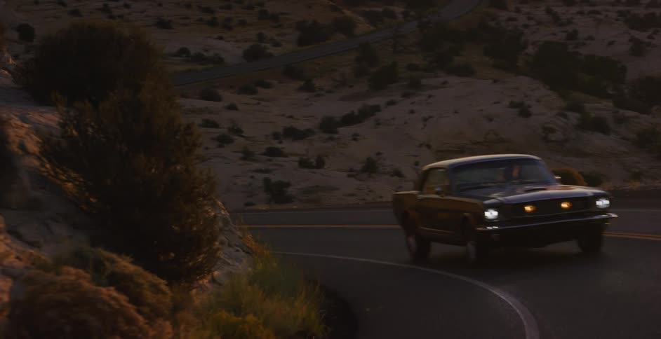 A classic black Mustang cruises down a scenic, winding desert highway at dawn, with the rising sun creating a beautiful lens flare