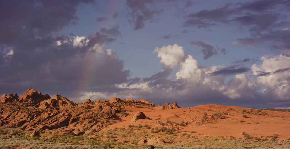 A vibrant rainbow arcs over a stunning red rock desert landscape under a dramatic, cloudy sky