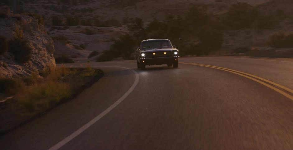 A vintage black muscle car with its headlights on drives along a winding desert road as the golden sunrise illuminates the landscape