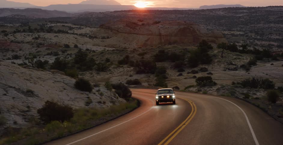 A vintage muscle car drives along a winding scenic route through a vast desert landscape at sunset
