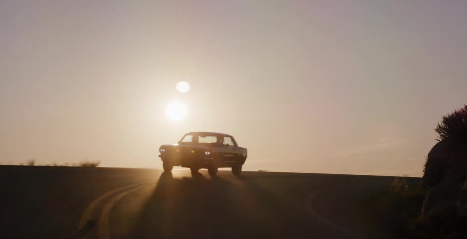 A vintage muscle car drives slowly on a winding road at dawn, silhouetted against the rising sun
