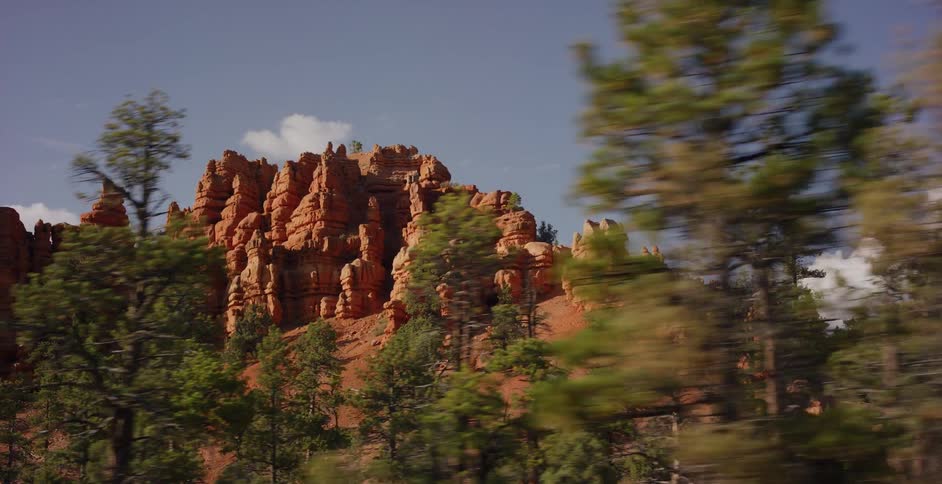 A scenic side-view driving shot reveals majestic red rock canyons and a lush pine forest on a sunny day