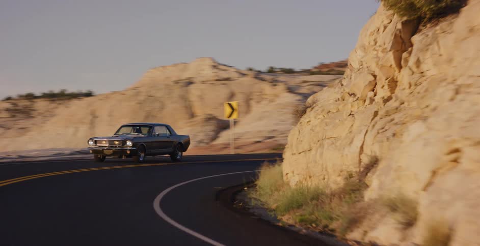 A vintage Ford Mustang cruises down a scenic highway through a rocky desert landscape during golden hour