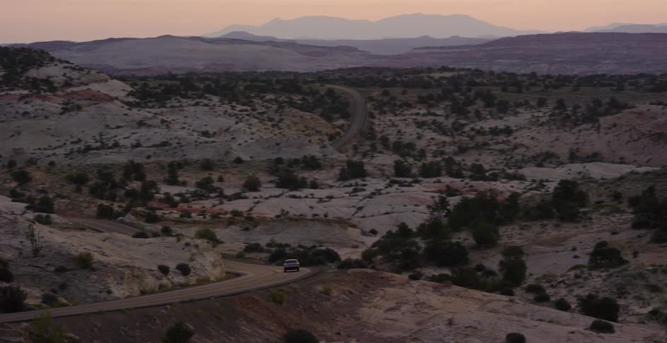 A solitary car drives along a winding scenic highway through a vast, rocky desert landscape at dusk