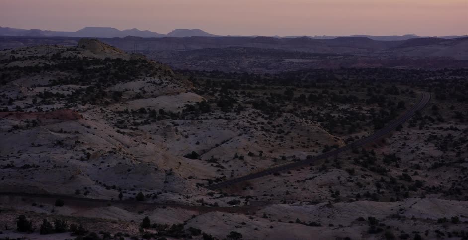 An aerial view shows a lone car driving along a winding road through a vast, rocky desert landscape at dusk