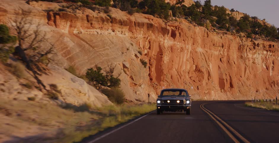 A classic black muscle car drives down a winding desert road next to red rock cliffs at golden hour