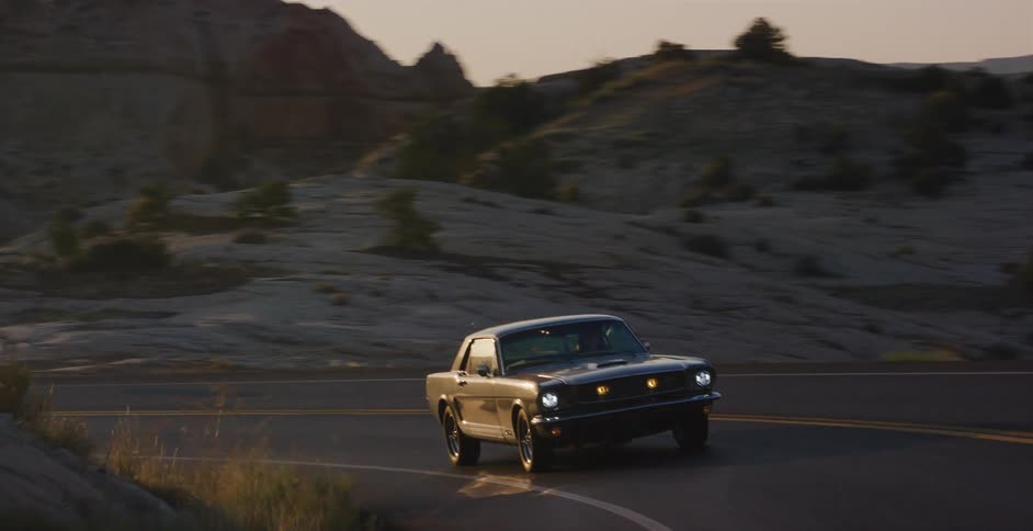 A classic black Ford Mustang with headlights on drives on a winding desert highway at sunset