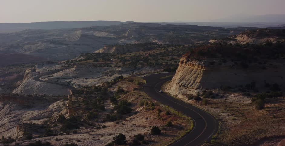 A stunning aerial view shows a lone vehicle driving on a winding scenic byway through a vast and rugged desert canyon at sunrise