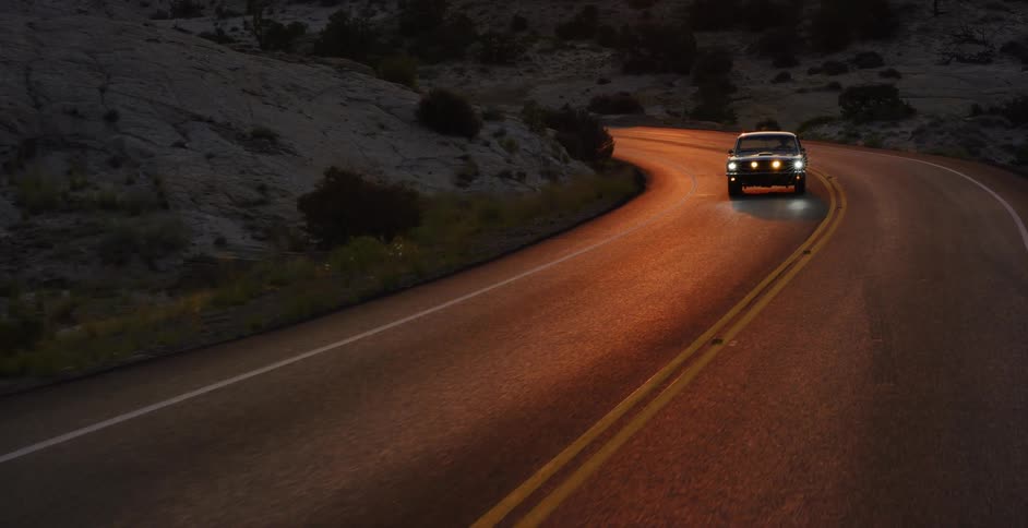 A classic black muscle car drives on a winding desert road at golden hour, the camera revealing the expansive landscape as the sun sets