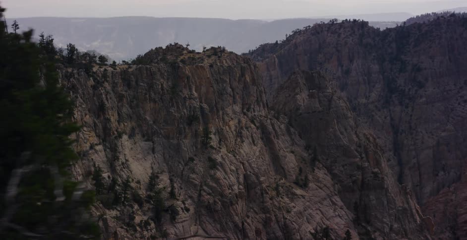 A slow-motion tracking shot past pine trees reveals a majestic and rugged mountain range under a hazy sky