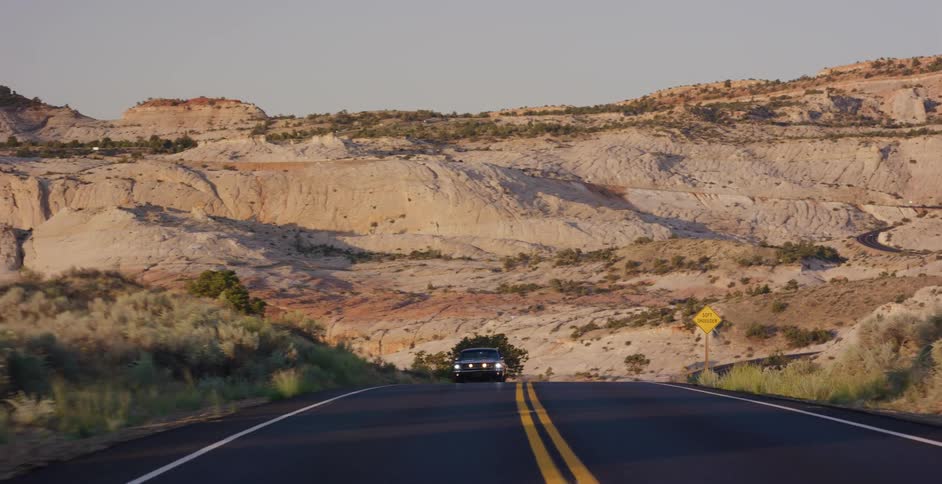 A classic black car with its headlights on drives towards the camera on a scenic highway winding through the vast, rocky desert landscape at dusk