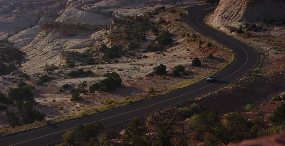 An aerial view shows a vintage car driving along a winding road through a vast and rocky desert landscape at sunrise