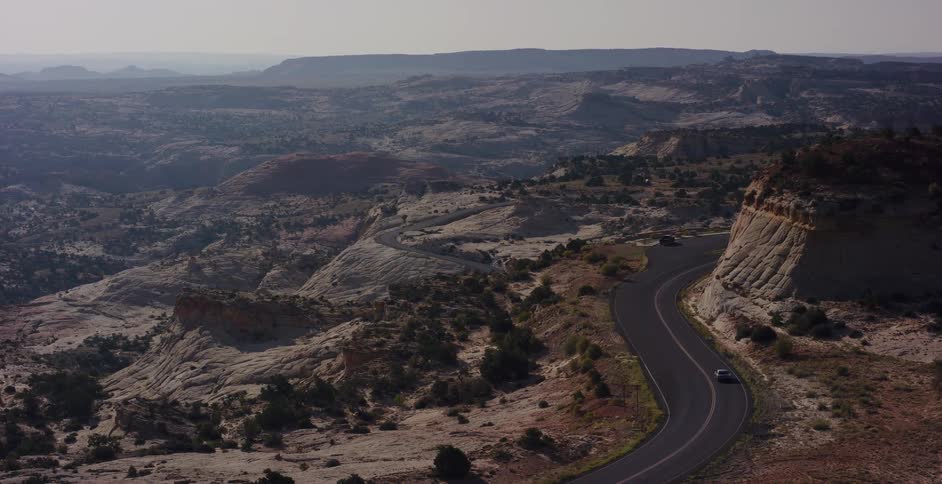 A car drives along a winding road through a vast, arid desert canyon landscape in a high-angle view