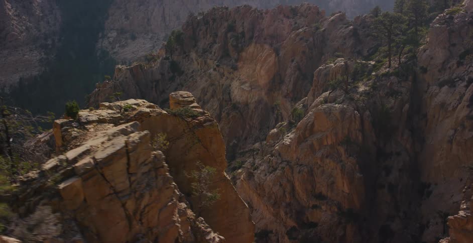 An expansive tracking shot moves along the edge of a deep, rugged canyon, revealing its vast, sunlit rock formations and distant, tree-covered mountains