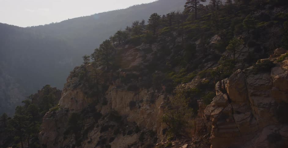 Sunlight filters through the haze over a rugged, forested mountain cliff in a slow pan shot