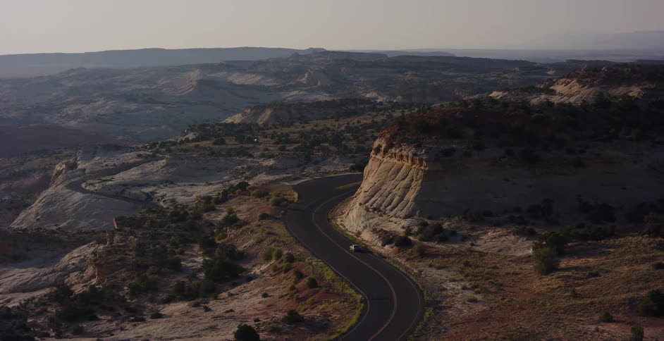 Aerial view of a solitary car driving on a winding scenic byway through a vast desert canyon landscape at sunrise