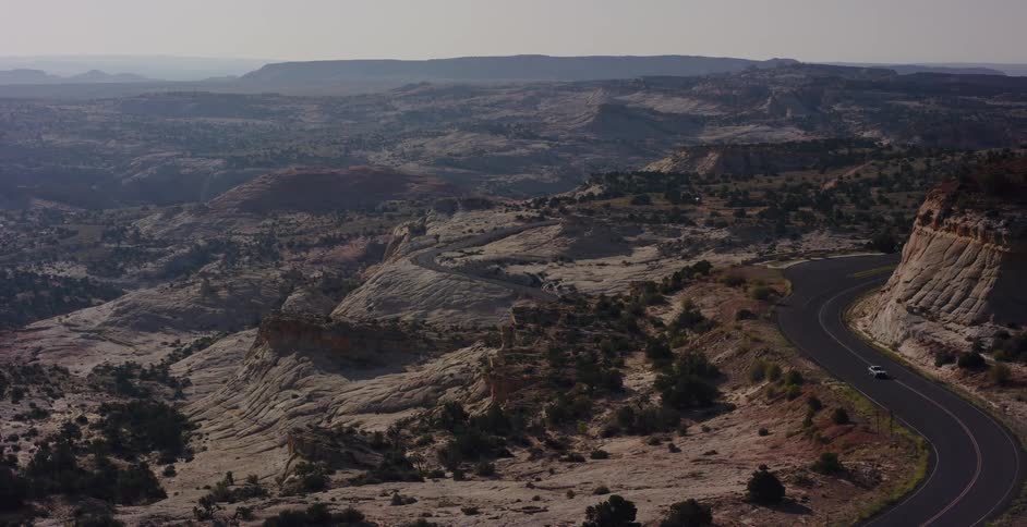 A lone car navigates a scenic winding road through a vast and hazy desert canyon landscape