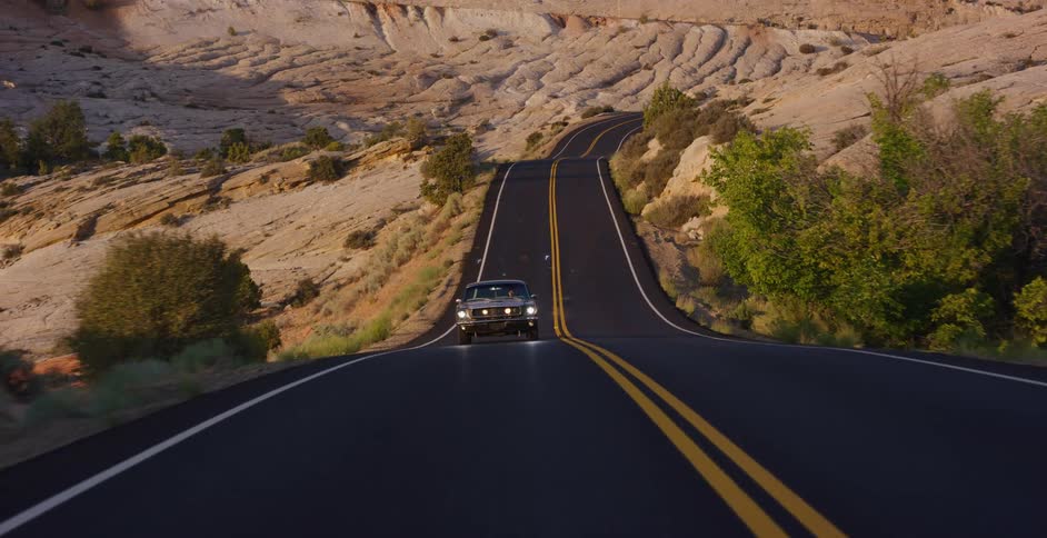 A classic black car drives along a scenic, winding road through a vast and beautiful desert landscape during the golden hour