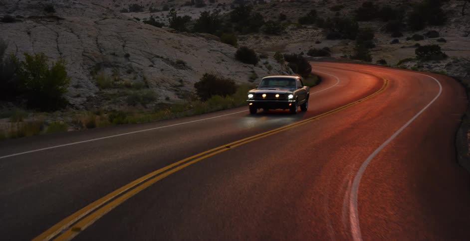 A classic Mustang drives along a winding desert highway as the camera reveals a breathtaking sunset over the mountainous landscape