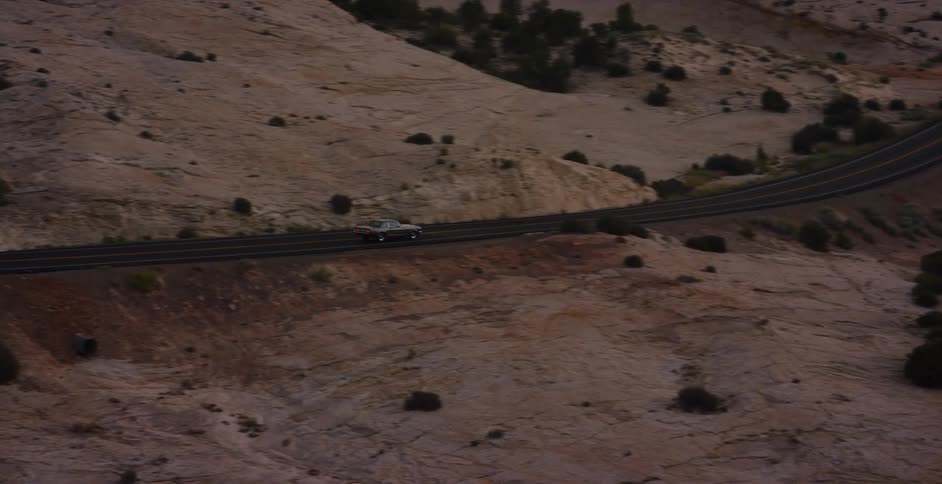 An aerial view shows cars driving along a winding highway through a vast and desolate rocky desert landscape at dusk