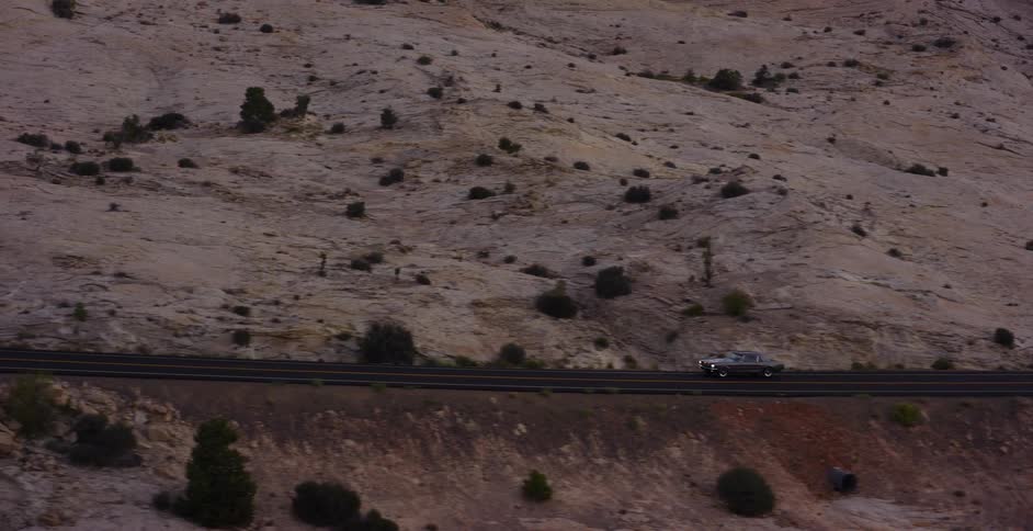 Aerial view of a vintage car cruising along a remote desert highway winding through majestic sandstone rock formations