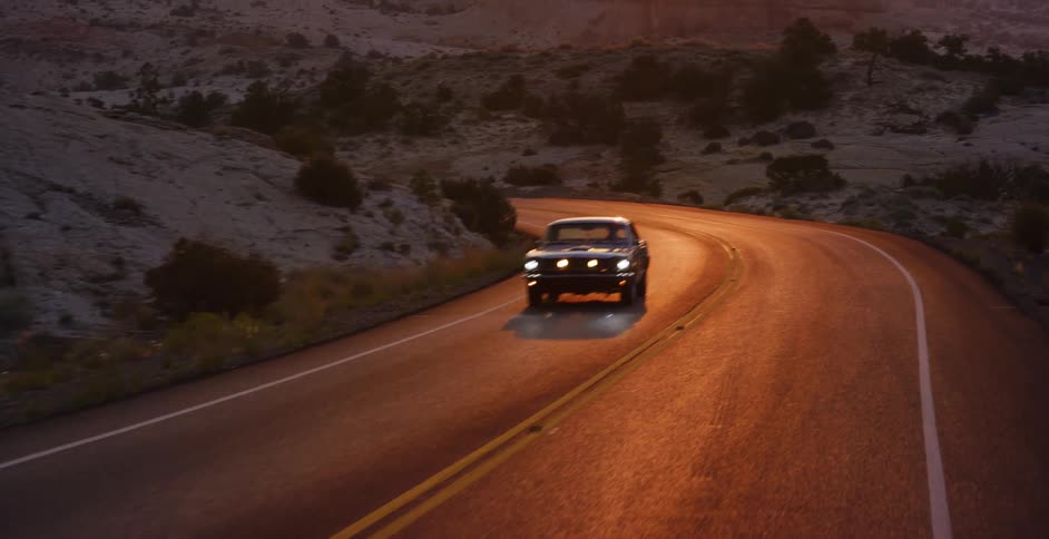 A classic black muscle car cruises down a winding desert highway as the camera reveals a stunning sunset over the canyons