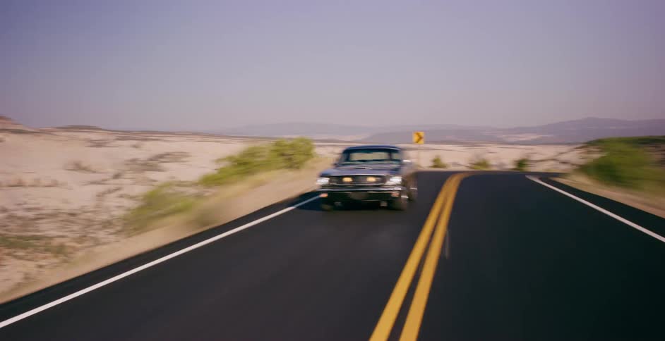 A classic black Ford Mustang speeds down a winding two-lane road through a scenic desert landscape