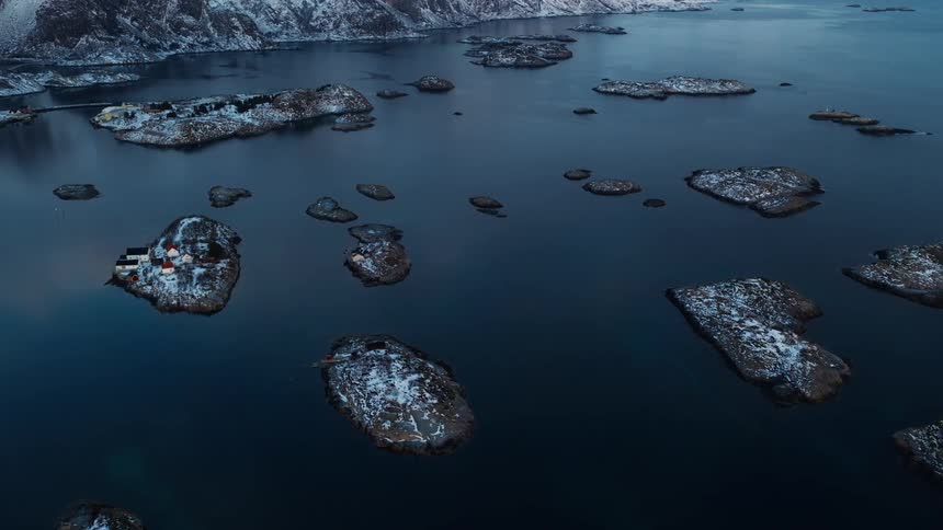 A stunning aerial view of a remote, snow-covered archipelago with small houses dotting the islands in a calm, dark blue fjord in Norway