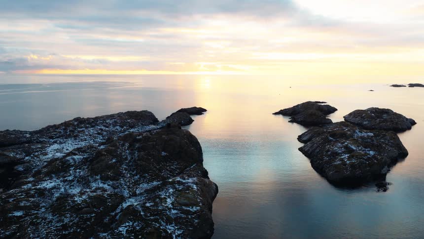 Serene aerial footage captures the tranquil beauty of snow-covered rocky islets reflecting in the calm sea during a golden sunrise