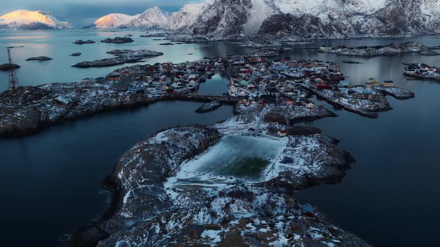 A stunning aerial pull-back reveals the snow-covered fishing village of Henningsvær in Norway's Lofoten Islands, showcasing its famous island football pitch against a backdrop of majestic mountains at sunrise