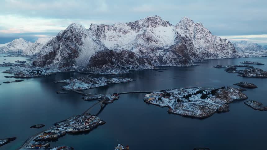 An epic aerial shot captures the stunning snow-covered mountains and island villages of Lofoten, Norway, during a calm winter twilight