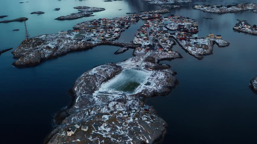 An aerial view pulls back from a snow-dusted soccer field to reveal the stunning Henningsvær village nestled among the Lofoten Islands in winter