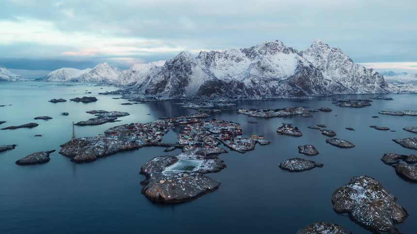 Static aerial view of a snowy fishing village on the Lofoten Islands in Norway, nestled among rocky islets with towering mountains in the background